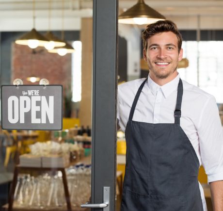 A white man is standing in the doorway of a restaurant with an open sign hanging in the door window.