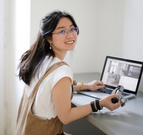 An Asian woman with glasses is sitting at a desk. She's holding a camera and working on a laptop.