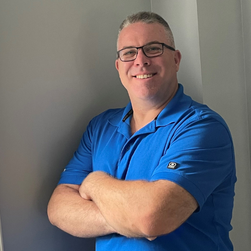 A white man smiling with short hair and glasses with his arms crossed