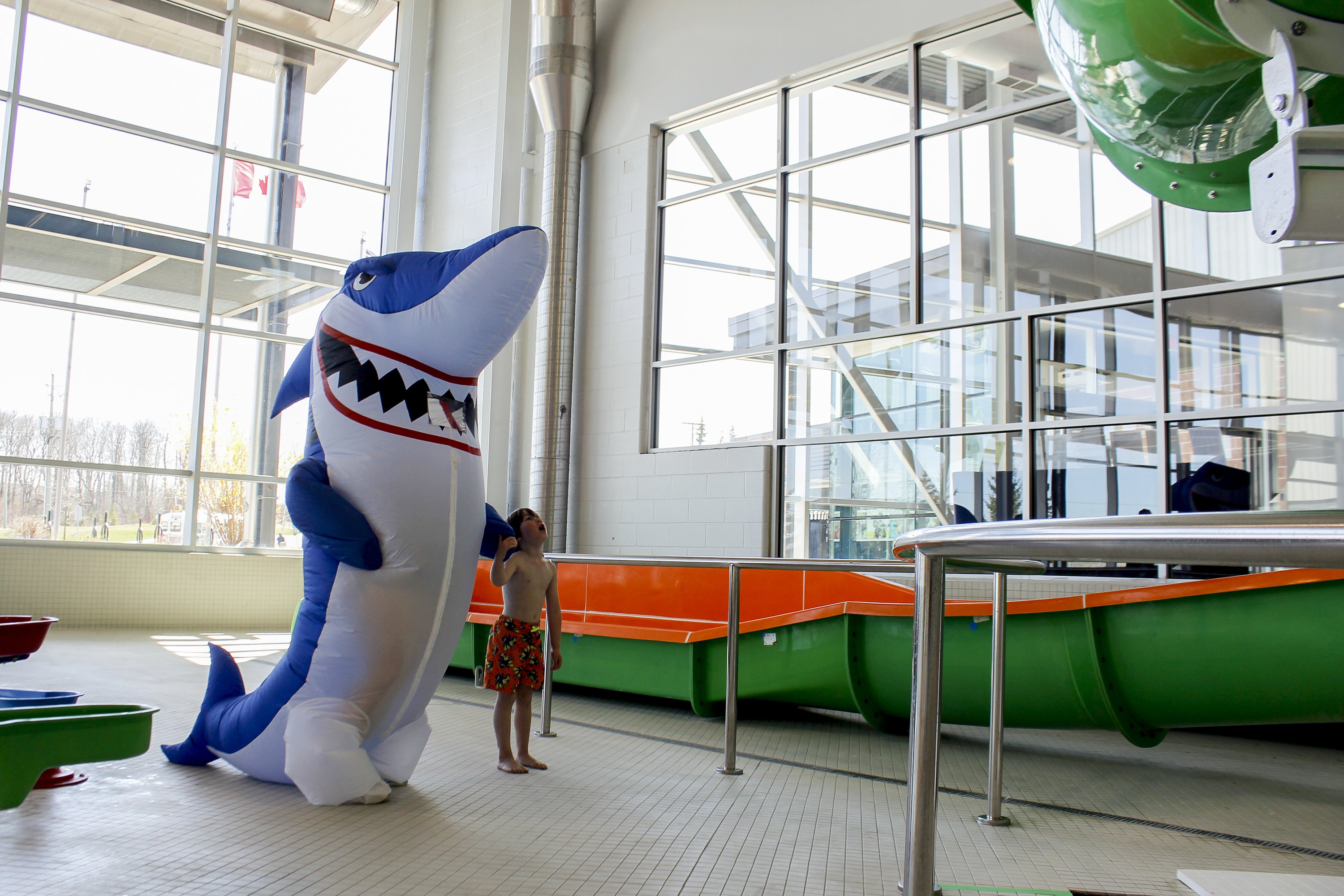 A Shark Costume standing beside a little boy staring up at the water slide